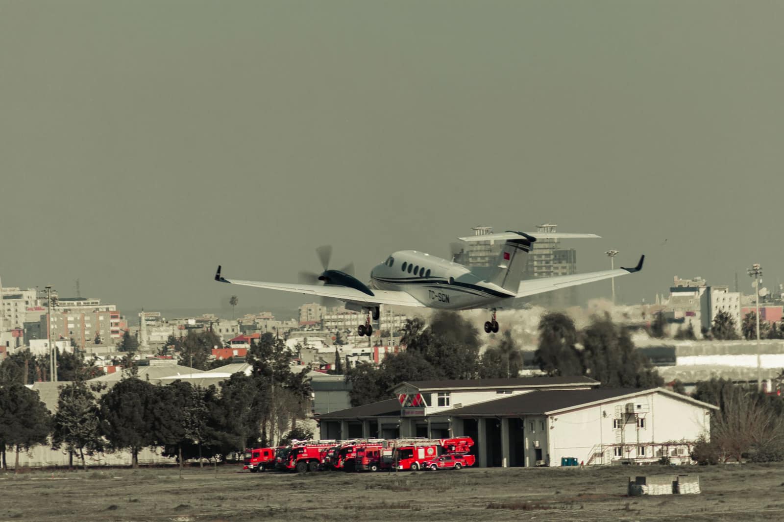 An airplane taking off over an urban skyline with airport fire trucks on standby below