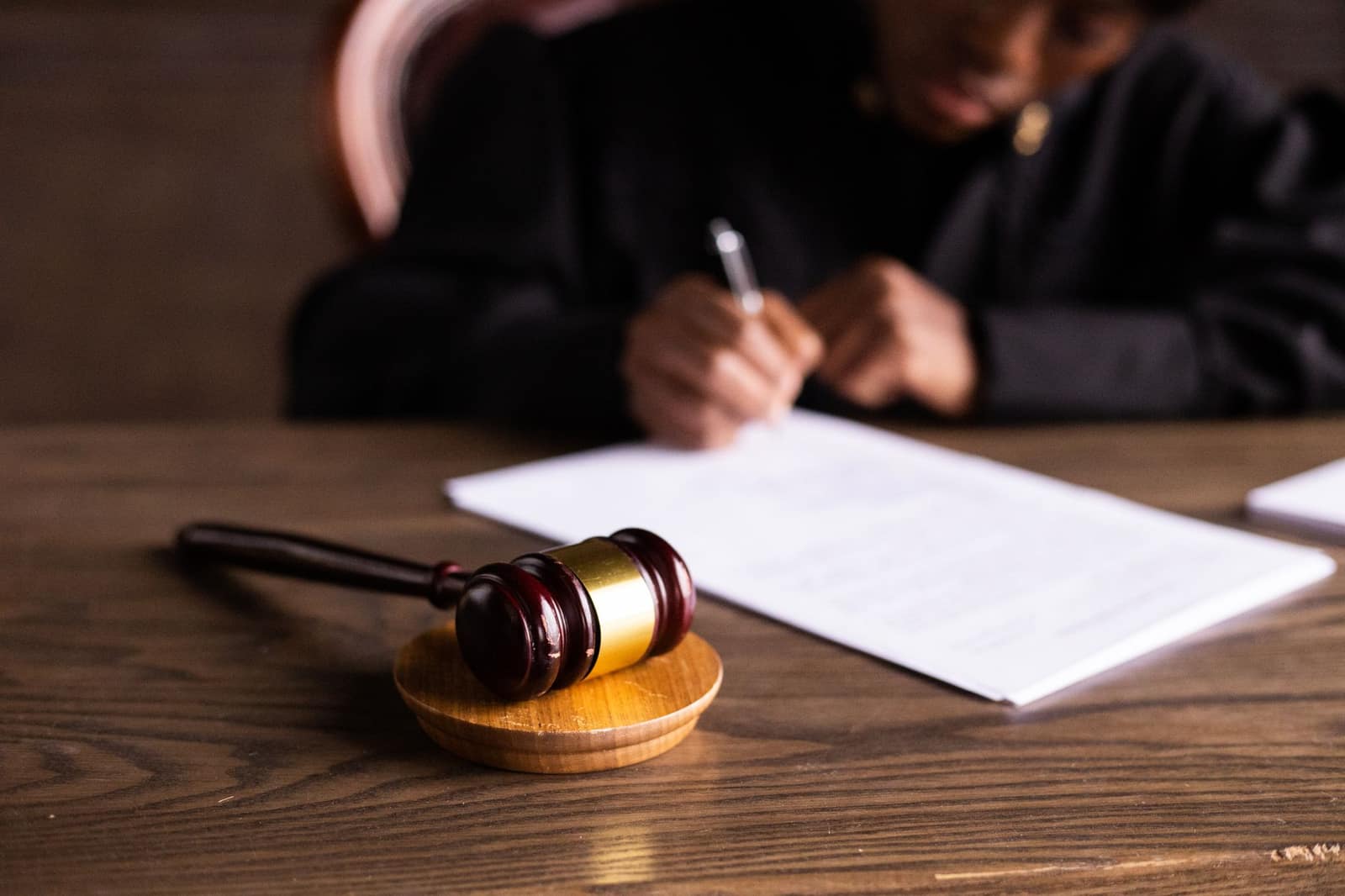 A gavel on a wooden sound block beside a judge signing documents at a desk