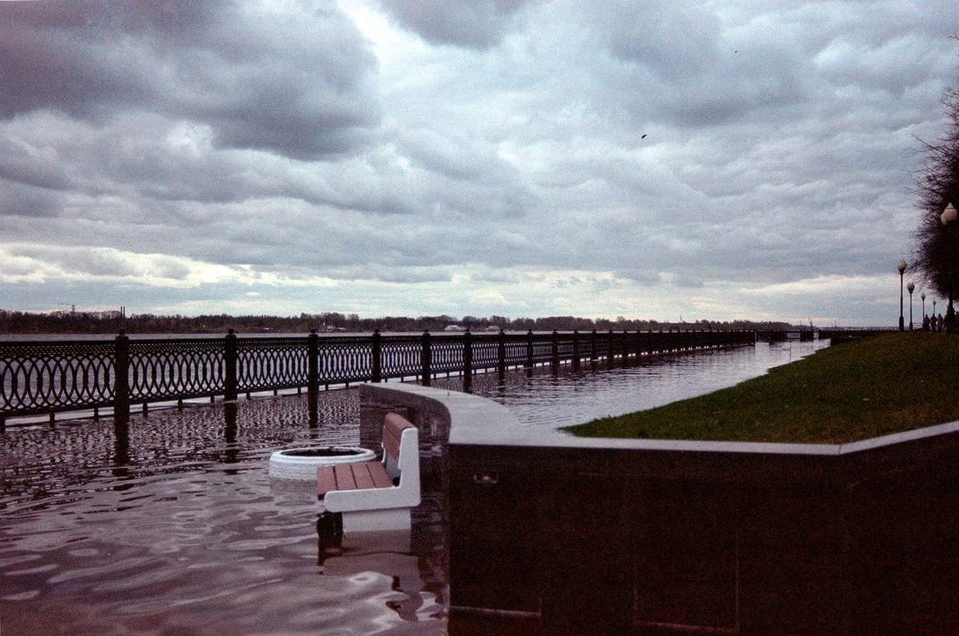 A flooded walkway along a riverbank with overcast skies and submerged benches