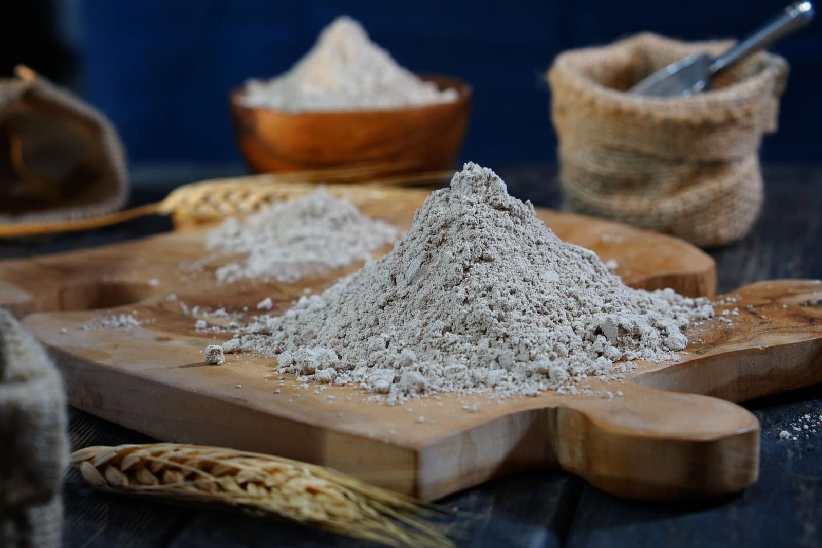 A pile of flour on a wooden board next to a sack and bowl of flour and stalks of wheat