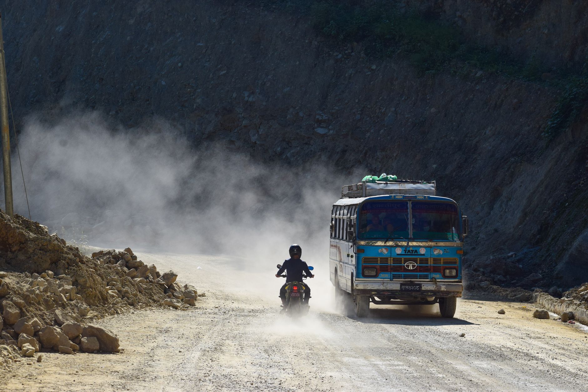 A bus and motorcycle on a dusty rural road surrounded by rocky terrain and rising dust clouds