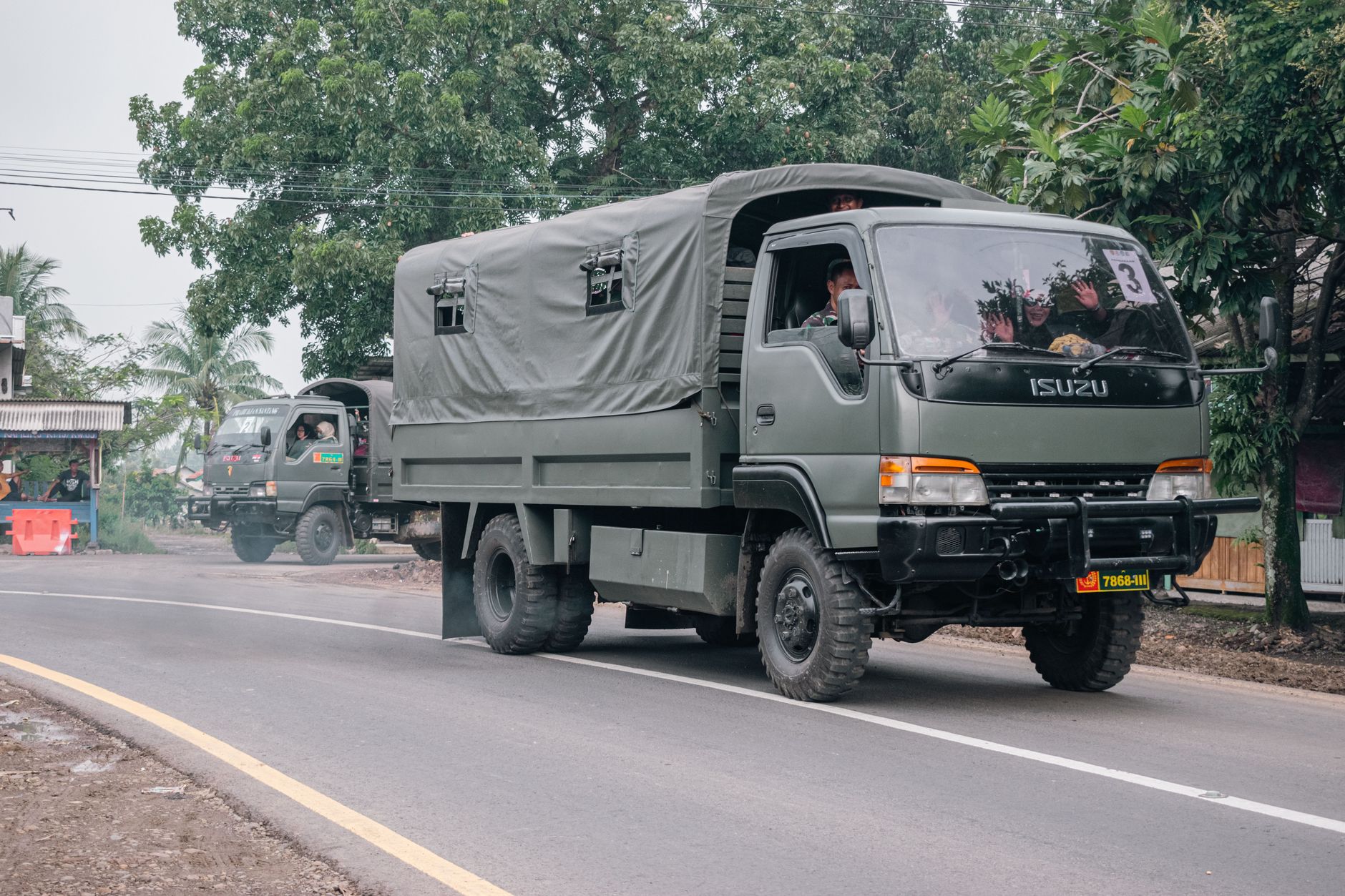Military trucks traveling on a rural road surrounded by trees and local buildings