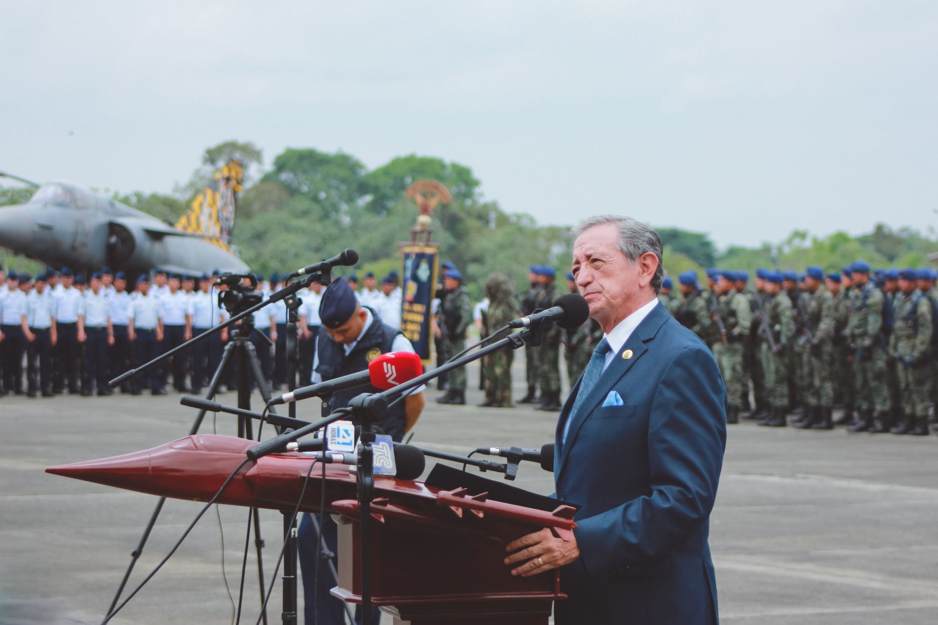 A military officer speaking at microphones during a press conference, with uniformed personnel and an aircraft in the background