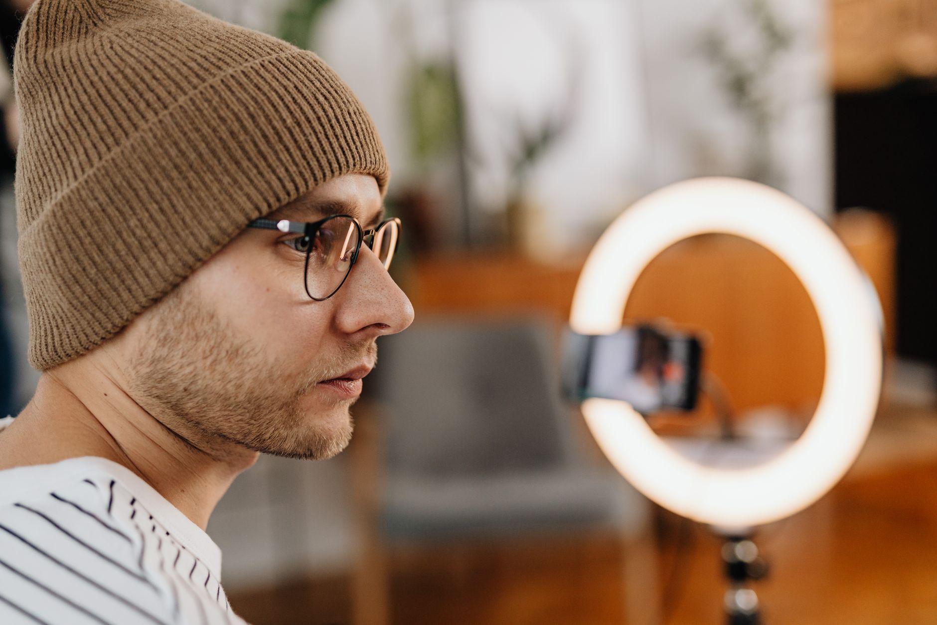 A man wearing eyeglasses and a knitted cap records a video at home using a smartphone and ring light