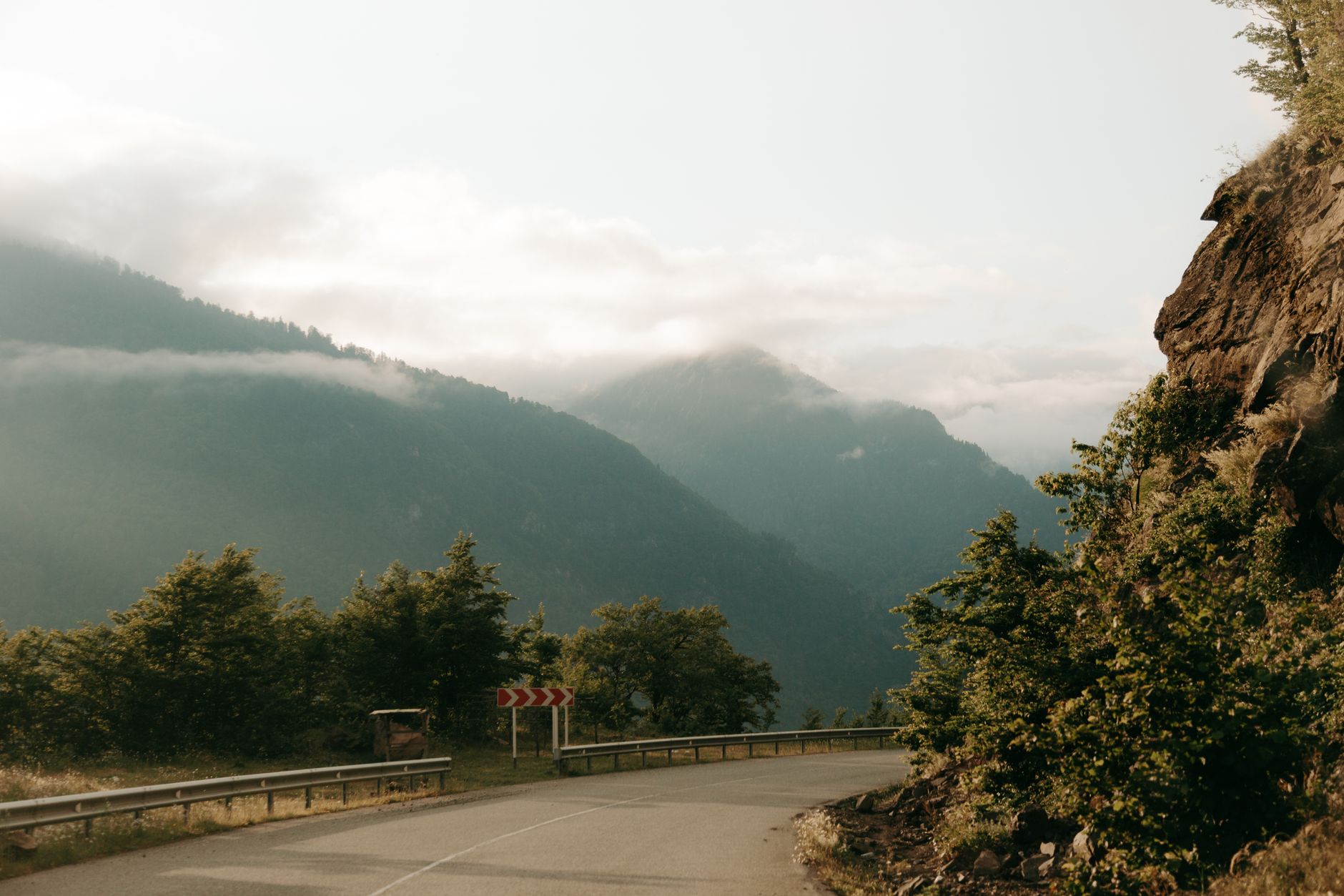 A scenic foggy mountain road winding through lush greenery with distant misty peaks