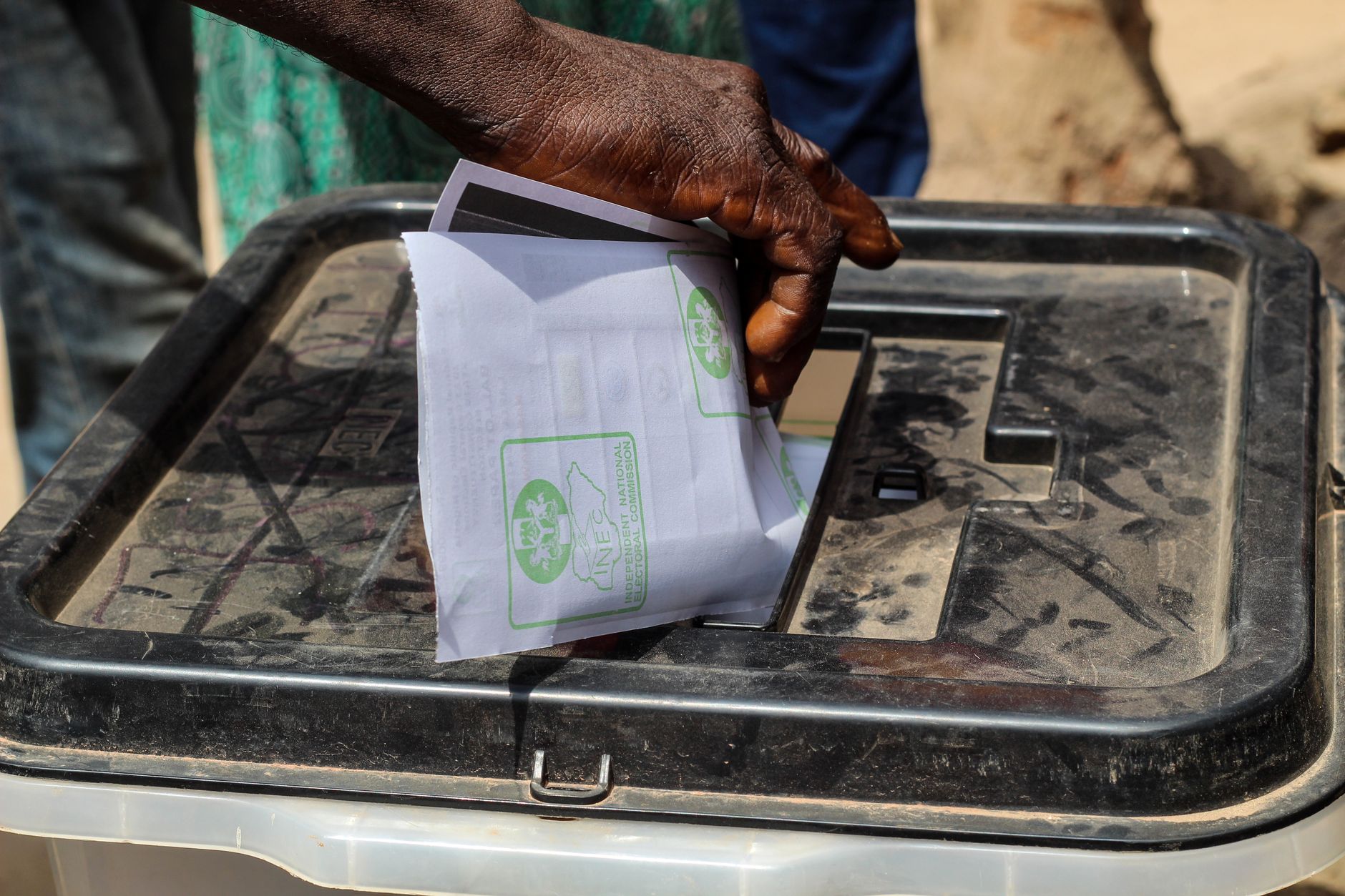 A hand placing a paper ballot into a transparent voting box outdoors