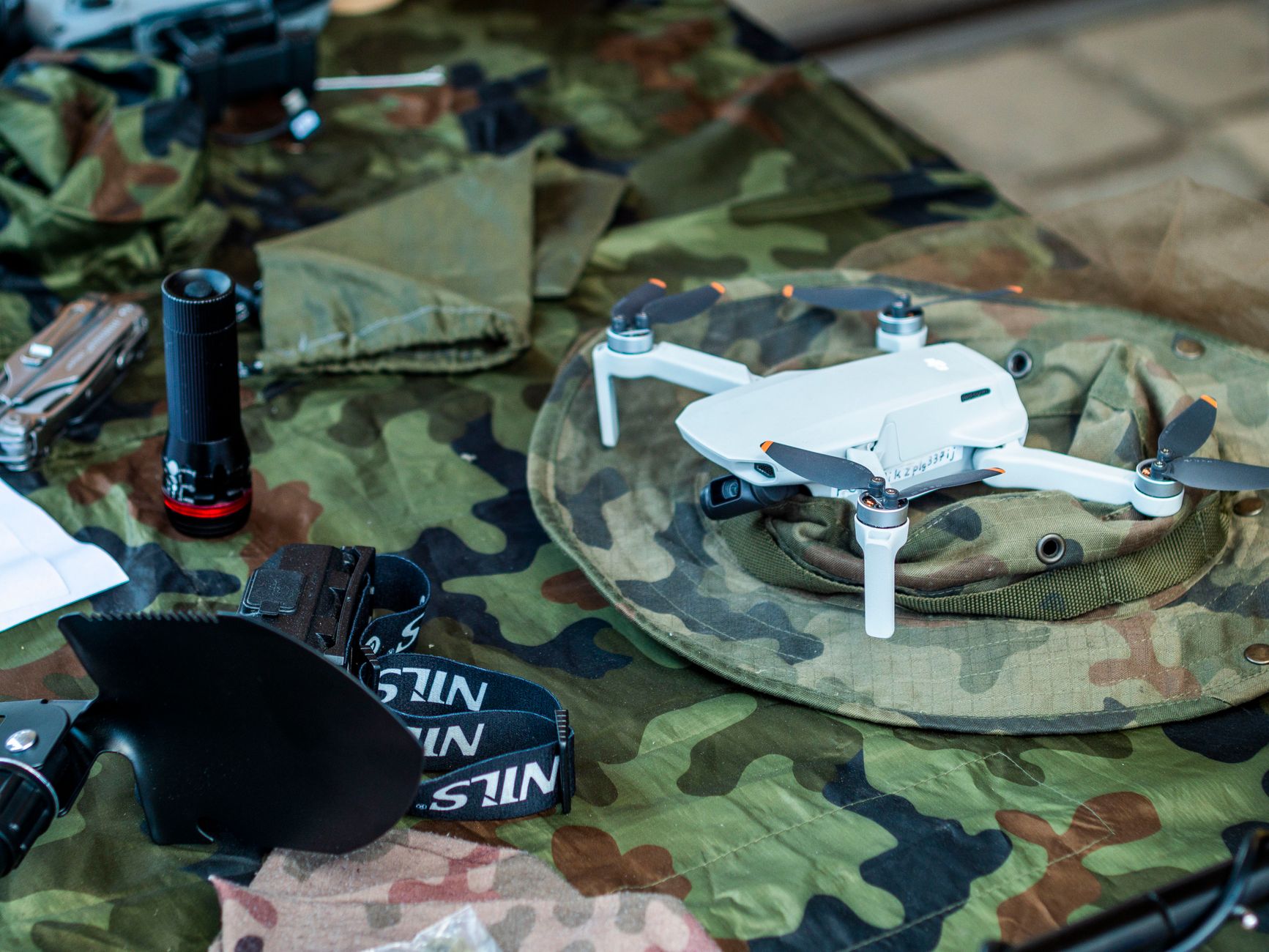 A small drone resting on camouflage military gear next to a flashlight and tools