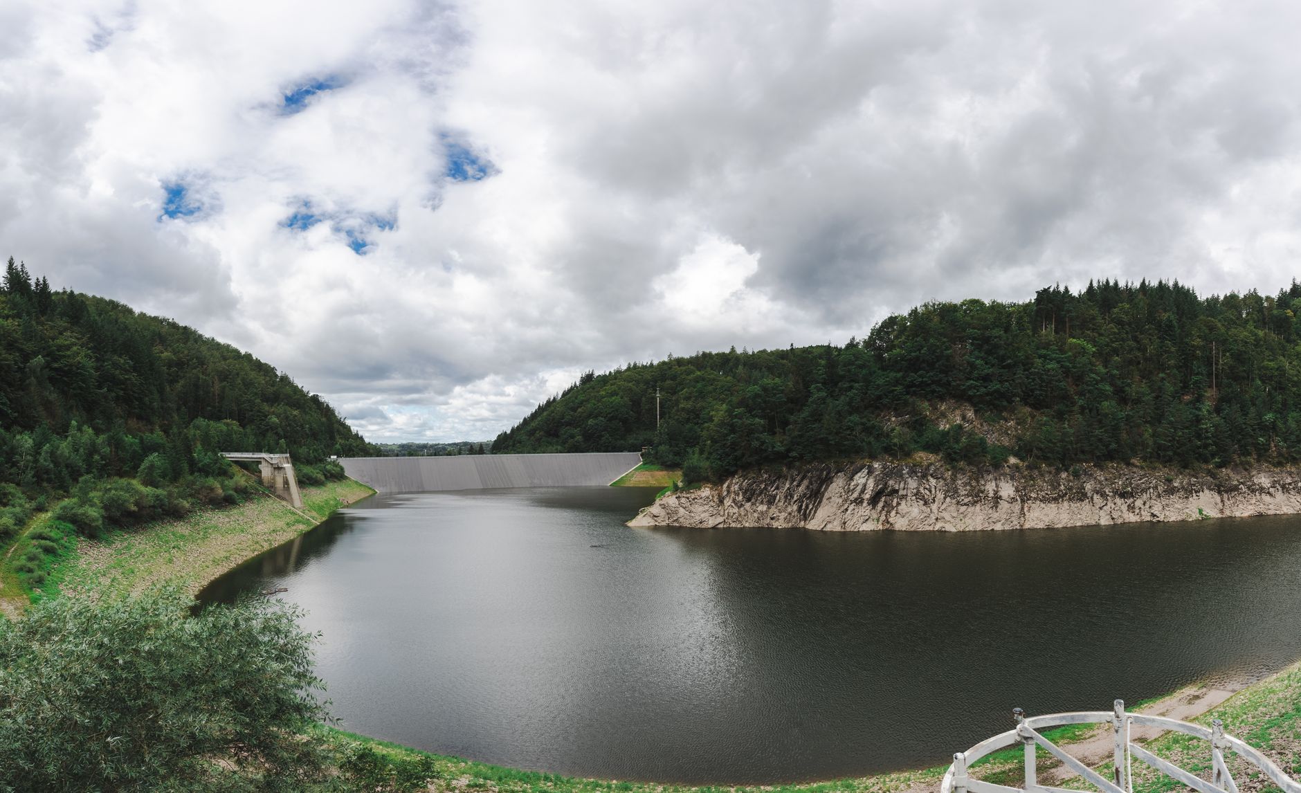 Panoramic view of a dam with a peaceful reservoir surrounded by lush green forests under a cloudy sky