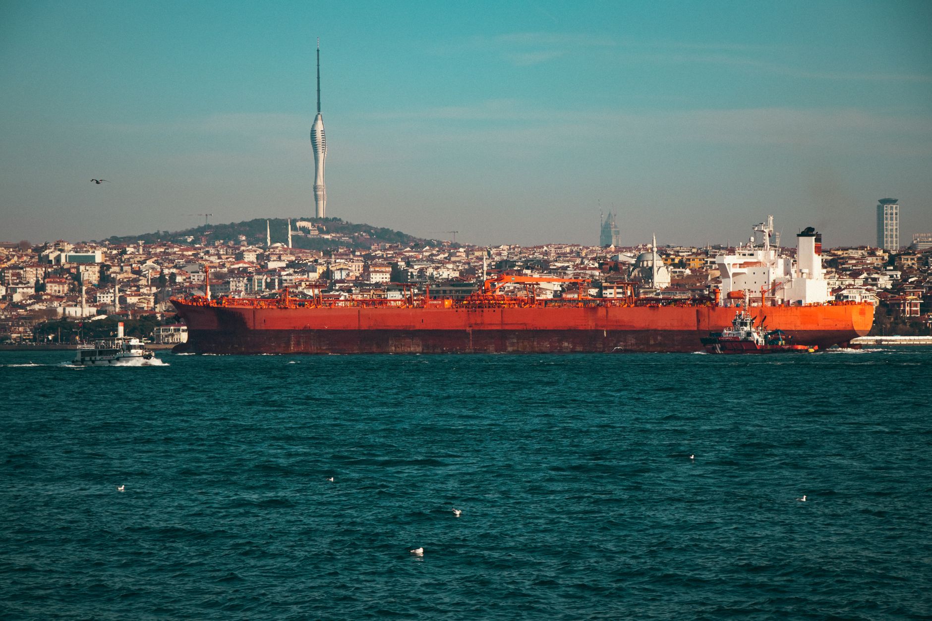 A large oil tanker navigates a strait waterway near a city skyline